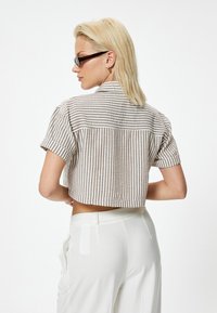 Camisa recortada con rayas verticales en marrón claro y blanco, mangas cortas y cuello de punta, combinada con pantalones blancos, mostrando la vista trasera.