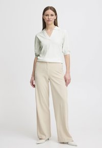 Blusa verde chiaro con scollo a V e maniche corte a sbuffo, abbinata a pantaloni beige a gamba larga e décolleté bianchi a punta.