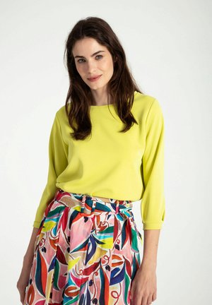 Bright yellow long-sleeve top paired with a colorful, patterned skirt featuring abstract leaves, belted at the waist. Smooth fabric texture.