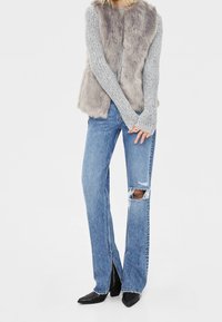 Faux fur grå väst över en texturerad grå tröja, tillsammans med ljusblå slitna jeans som har ett hål vid knät och svarta spetsiga stövlar.