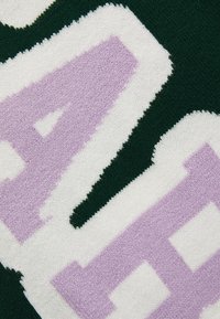 Strickstoff mit großen, strukturierten Zahlen in Lila und Weiß, umrandet von Grün. Weiche, flauschige Textur auf einem dunkelgrünen Hintergrund.