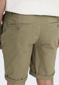 Shorts en coton kaki avec une finition texturée, présentant des ourlets roulés, deux poches arrière avec des boutons et des détails de couture.