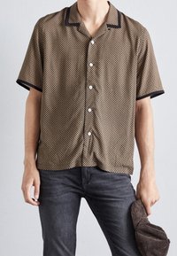 Korte mouwen, bruine shirt met patroon en een zwarte kraag, voorzien van knopen aan de voorkant en een relaxte pasvorm. Gecombineerd met donkere jeans en een hoed.