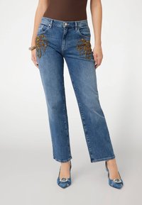 Šviesiai mėlyni džinsai su tiesiomis kojomis, turintys sudėtingus karoliukų raštus priekyje ant kišenių. Suderinti su mėlynais dekoruotais aukštakulniais.