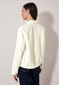 Witte denim jas met een kraag, knopenmanchetten en een rechte snit. Achterzijde bevat een horizontale naad en zijsplitten. Draagt over donkere jeans.