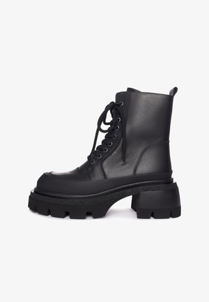 Bota de combate de cuero negro con suela gruesa, cordones en la parte frontal, tobillo alto y asa en el talón sobre fondo blanco.