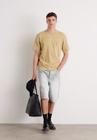Beige, rövid ujjú ing, cipzáras zsebbe, világos csíkosSorttal párosítva. A modell egy fekete kézitáskát tart, és fekete zoknit és cipőt visel.