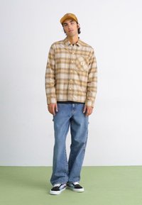 Rutete beige og brun skjorte med knapper, lyseblå vide jeans og svarte, hvite og grønne sneakers. Brukt med en tan cap.