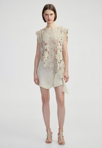 Haut sans manches beige clair avec un motif en dentelle intricate et des bords festonnés, associé à une jupe crème drapée. Sandales à talons beige à lanières.