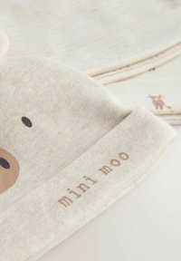 Gorro de algodón color beige claro con parte superior redondeada, que presenta un patrón minimalista de cara de vaca y el texto "mini moo" en marrón suave en el puño.