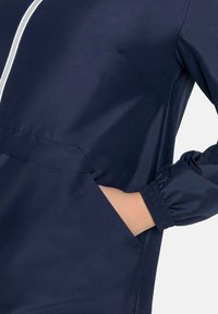 Giacca a vento navy con tasca frontale, polsini elasticizzati e colletto alto. Caratterizzata da una cerniera bianca che attraversa la parte anteriore per la chiusura.