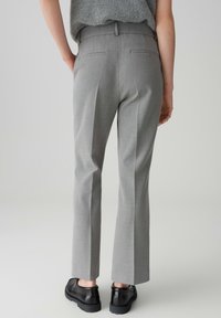 Persona che indossa pantaloni grigi su misura e mocassini neri, visto da dietro su uno sfondo grigio uniforme.