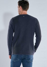 Marineblauer Langarmpullover aus strukturiertem Stoff, Rundhalsausschnitt und Etikett auf der Rückseite, kombiniert mit blauen Jeans.