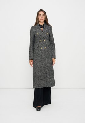 Femme debout portant un long manteau croisé gris à chevrons avec des boutons dorés et un pantalon noir à jambes larges sur un fond blanc uni.