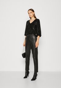 Schwarzes Pailletten-Top mit V-Ausschnitt und Dreiviertelärmeln, kombiniert mit schwarzen Lederhosen und schwarzen ankle boots mit Absatz. Schwarze Tasche mit Kettenriemen.
