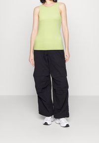 Persona che indossa un top senza maniche verde chiaro, pantaloni cargo neri larghi e sneakers bianche, in piedi davanti a uno sfondo bianco semplice.