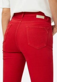 Pantaloni in denim rosso con design aderente, vita media e due tasche posteriori. Presentano dettagli di cucitura sottili vicino alle tasche.