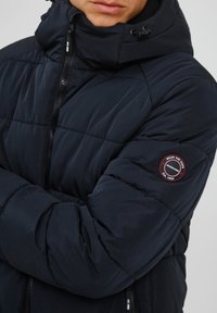 Mornarska puffer jakna s visokim ovratnikom, kvadratnim uzorkom i okruglom logotipom na lijevoj ruci. Ima prednji patentni zatvarač i podesivu kapu.