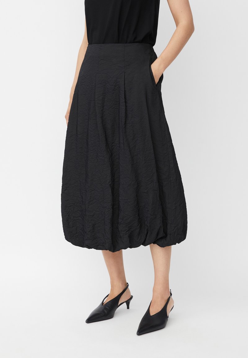 Donna che indossa una gonna a palloncino nera midi con texture e tasche, insieme a scarpe con tacco a punta nere e a schiacciare, su uno sfondo neutro.