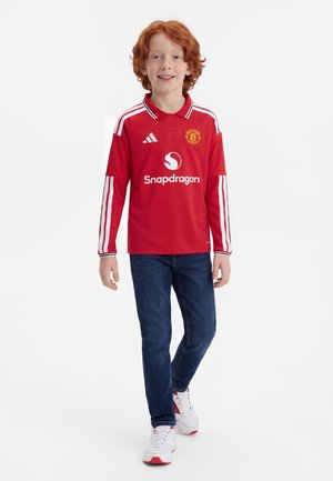 Rudy chłopiec w czerwonym, długim podkoszulku Manchester United, ciemnoniebieskich dżinsach i białych trampkach, idzie naprzód na białym tle.