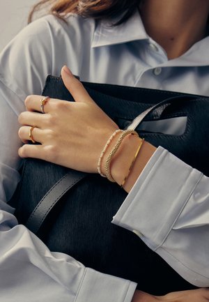 Valge särgiga naine hoiab musta käekotti, kandes vasakul käel kuldseid ja pärlikeega käevõrusid ning kuldseid sõrmuseid.