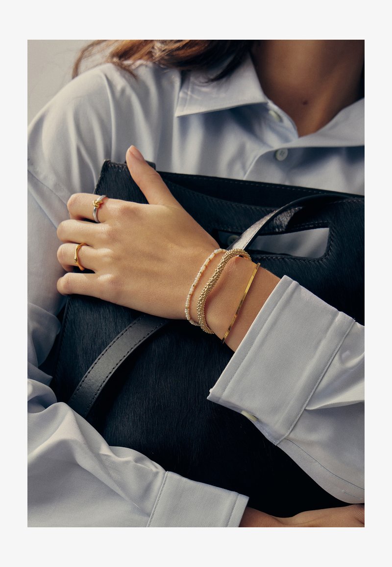 Γυναίκα με λευκό πουκάμισο που κρατάει μαύρη τσάντα, φοράει χρυσά και περλέ βραχιόλια και χρυσά δαχτυλίδια στο αριστερό χέρι.