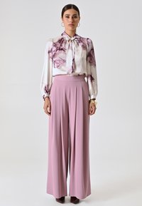 Blusa bianca con stampa floreale viola, scollatura alta e fiocco. Pantaloni rosa a gamba larga con pieghe. Orecchini rotondi semplici.