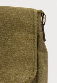 Τσάντα ώμου ελιάς με υφή υφάσματος, με κλείσιμο με καπάκι και μεταλλικό κούμπωμα. Ορατή ραφή κατά μήκος των άκρων.