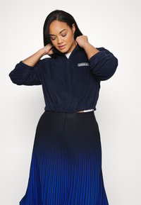 Πολυθρόνα φλις ναυτικού μπλε με σχέδιο cropped και ελαστική μέση, συνδυασμένη με φούστα πλισέ σε γραδαριστό σχέδιο που μεταβαίνει από το μαύρο στο μπλε.