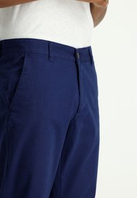 Pantalones azul marino confeccionados en una mezcla de algodón con un corte ajustado, que cuentan con un bolsillo frontal, costuras laterales y un cierre de botón.