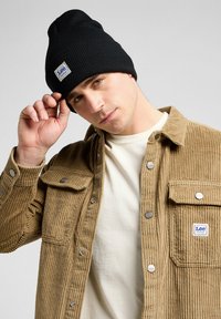 Zwarte ribgebreide beanie gedragen met een tan corduroy jas met een knoopsluiting, twee borstzakken en een klein logo-opdruk op de zak.