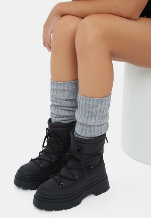 Bottes de cheville noires imperméables avec semelles épaisses et design à lacets. Associées à des chauffe-jambes en tricot côtelé gris roulés pour plus de chaleur.