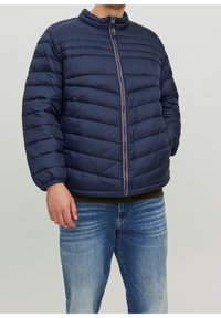 Marineblaue Steppjacke mit horizontalem Steppmuster, Stehkragen und vorderem Reißverschluss. Kombiniert mit einem grünen Oberteil und hellblauen Jeans.