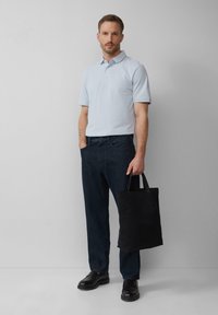 Hellblaues Poloshirt, dunkelblaue Jeans und schwarze Slippers. Hält eine schwarze Tragetasche. Einfaches Design, lässige Passform und dezente Textur.