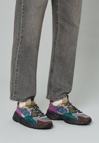Sneaker multicolori caratterizzati da una combinazione di materiali grigi, verdi, viola e marroni, con una suola nera texturizzata e lacci a contrasto.