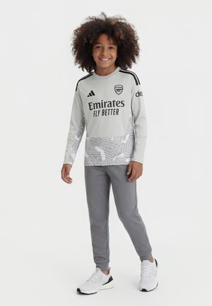 Enfant souriant aux cheveux bouclés portant un maillot de football gris d'Arsenal, pantalon assorti et baskets blanches, debout sur un fond neutre.