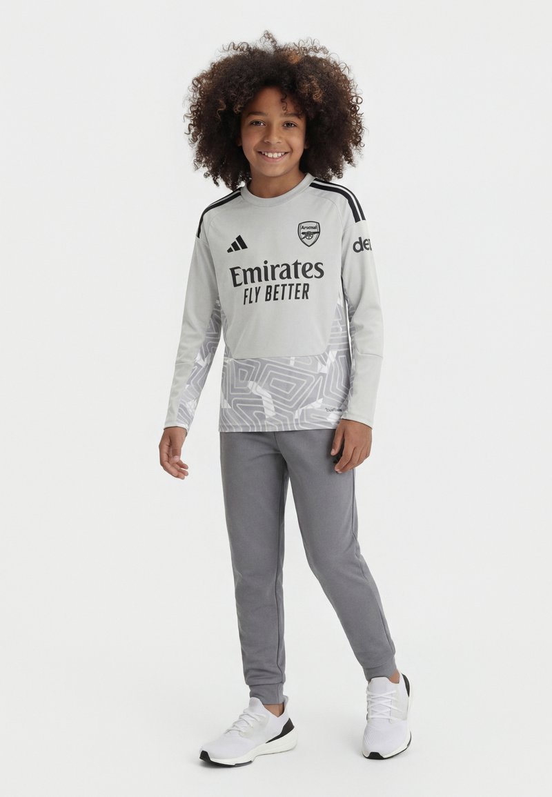 Enfant souriant aux cheveux bouclés portant un maillot de football gris d'Arsenal, pantalon assorti et baskets blanches, debout sur un fond neutre.