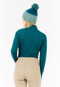 Tyrkysový svetr s dlouhým rukávem a vysokým límcem je kombinován se světlými béžovými kalhotami. Modelka má na hlavě pletenou čepici v tyrkysové a světle modré barvě s bambulkou.