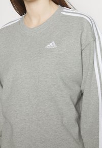 Närbild av en person som bär en grå Adidas-tröja med vita ränder på ärmarna och en vit Adidas-logga på bröstet.