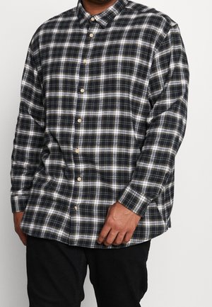 Mand iført en sort og hvid ternet skjorte med lange ærmer og sorte bukser, stående foran en ensfarvet baggrund.