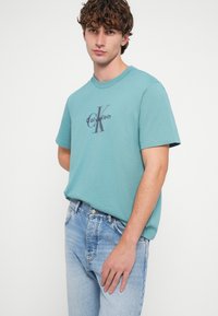 Šviesiai žaliai mėlynas medvilninis marškinėlis trumpomis rankovėmis su tamsiu grafiniu logotipo spaudiniu. Derinamas su išblukusiais mėlynais džinsais.