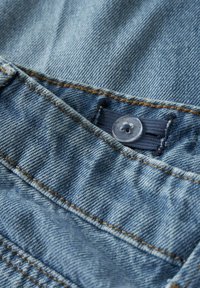 Lys blå denimstoff med en synlig knapp og søm. Den teksturerte overflaten viser et tradisjonelt vevmønster med lommer.