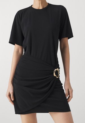 Kvinde iført en kort sort kjole med korte ærmer, rynket stof og en dekorativ guldbeklædning på højre side ved taljen.