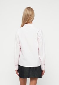 Mulher com cabelo longo loiro, vestindo uma camisa de manga longa rosa clara e uma saia cinza escura, de costas contra um fundo branco liso.