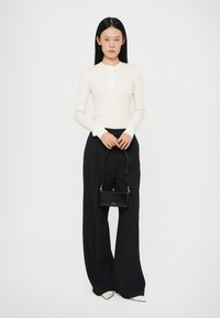 Top a maniche lunghe di colore crema con patta a bottoni, abbinato a un pantalone wide leg a righe nere e borsa nera piccola con logo dorato.