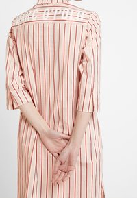 Vestido de camisa a rayas, de color rosa claro con líneas verticales rojas. Tiene un cuello con solapa, mangas tres cuartos y un logotipo en la parte posterior.