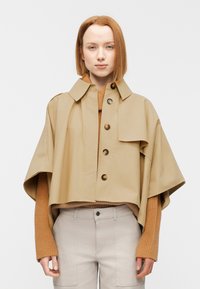 Veste beige courte avec un col à revers, fermeture à boutons et larges manches. Portée sur un pull côtelé couleur terre cuite et un pantalon gris clair.