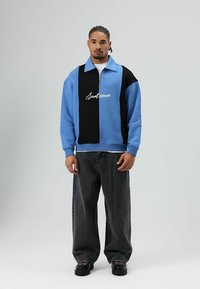 Sweat-shirt bicolore bleu et noir avec col zippé et logo imprimé à l'avant, associé à un jean noir ample et des chaussures noires.