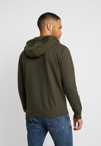 Olivgrüner Pullover mit Zugkordelkapuze, gerippten Bündchen und Saum, kombiniert mit blauen Jeans, die eine detaillierte Steppnaht an den Gesäßtaschen aufweisen.