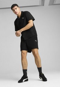 Čierny polo tričko s krátkym rukávom a ladiace šortky, textúrovaná látka, s bielym logom Puma. Nosené s čiernymi teniskami a ponožkami, na jednofarebnom pozadí.